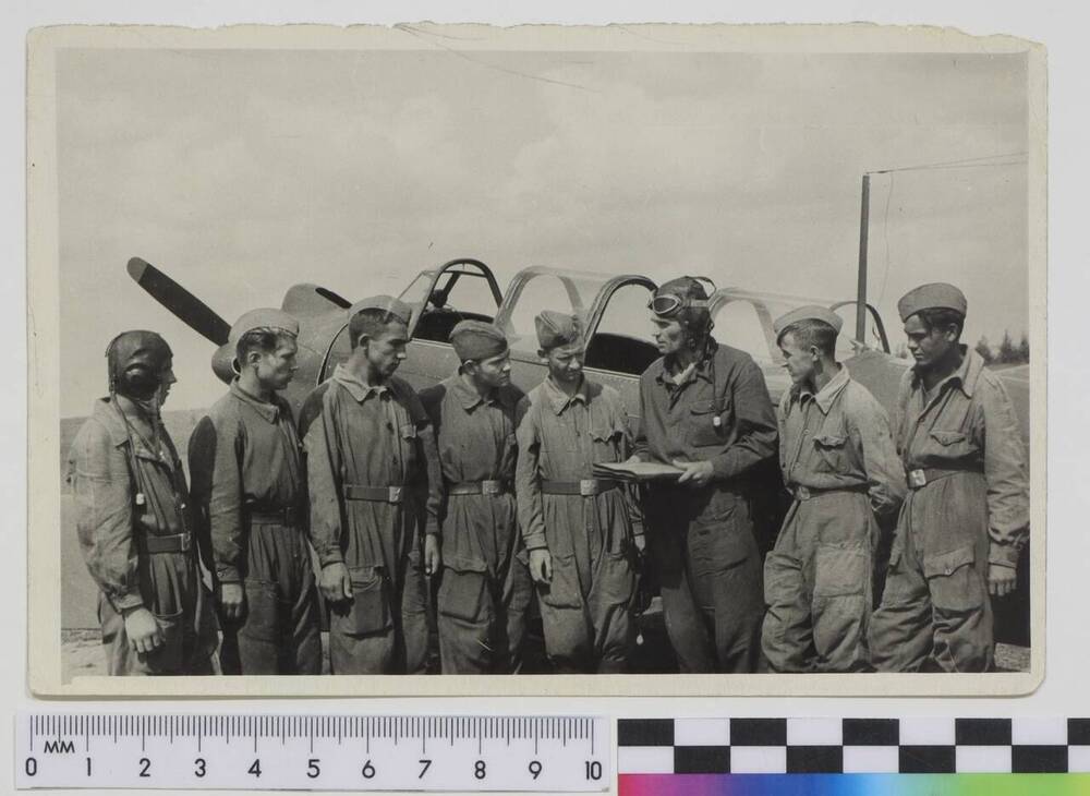 Фото групповое покаленное на фоне учебного самолёта. Панкратов А.А. в шлемофоне 3-й справа проводит занятия с курсантами аэроклуба 1960-е гг.
