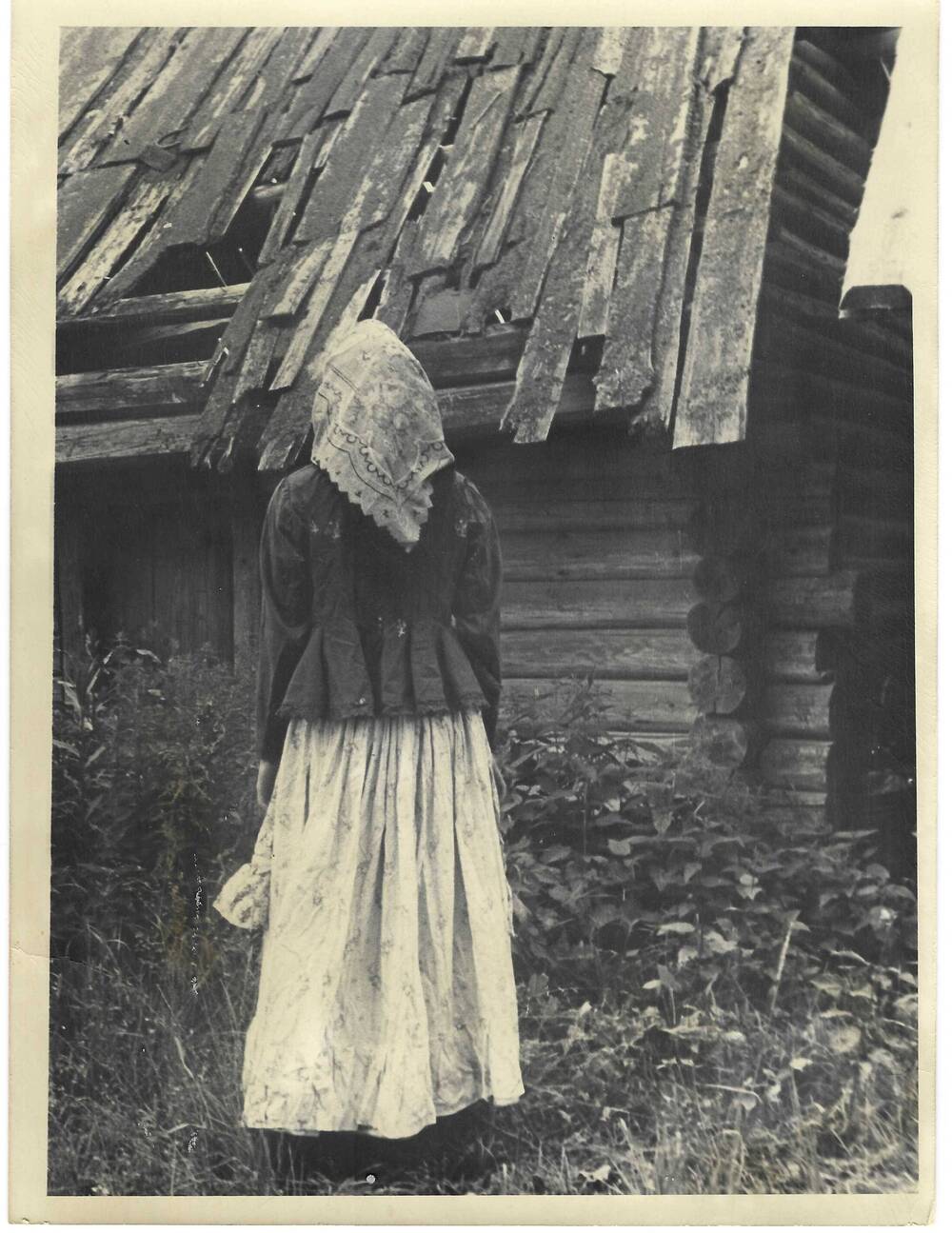 Фото. Ростовой портрет женщины в традиционном костюме на фоне деревянной хозяйственной постройки.