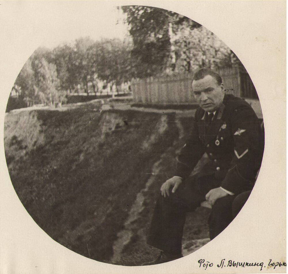 В.П. Чкалов в военной форме, с орденами, без головного убора, сидит на скамейке, на крутом берегу Волги около забора родного дома.