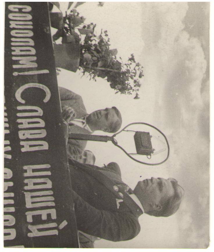 В.П. Чкалов в штатском костюме с орденами , с открытой головой  стоит на трибуне  перед микрофоном и рассказывает трудящимся г. Горького о своем перелете.