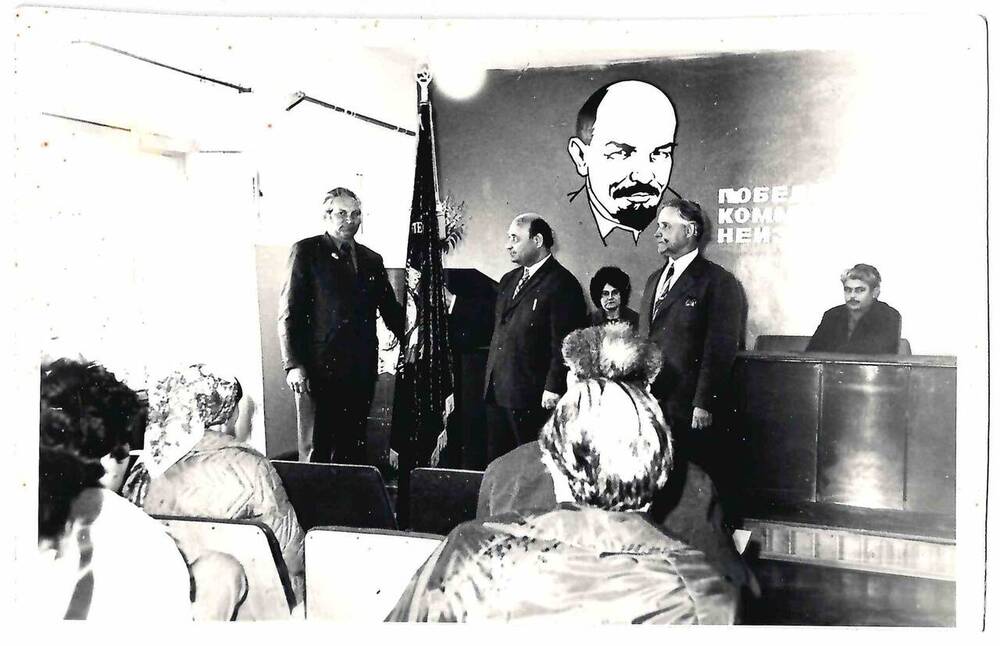 Фотография черно-белая событийная. Запечатлено мероприятие в актовом зале. Справа от знамени Саенко Ф.А. 