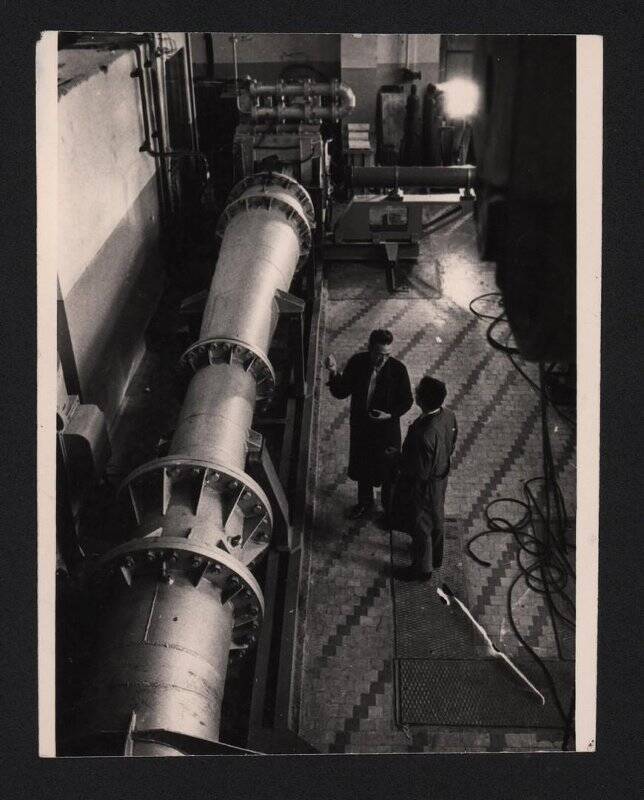 Фотография черно-белая. Двое мужчин в темных халатах стоят возле прибора в виде металлической горизонтальной трубы.