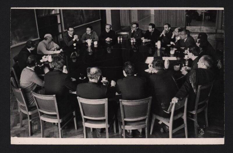 Фотография черно-белая. Группа ученых сидит за большим круглым столом черного цвета в Институте ядерной физики СО АН СССР.