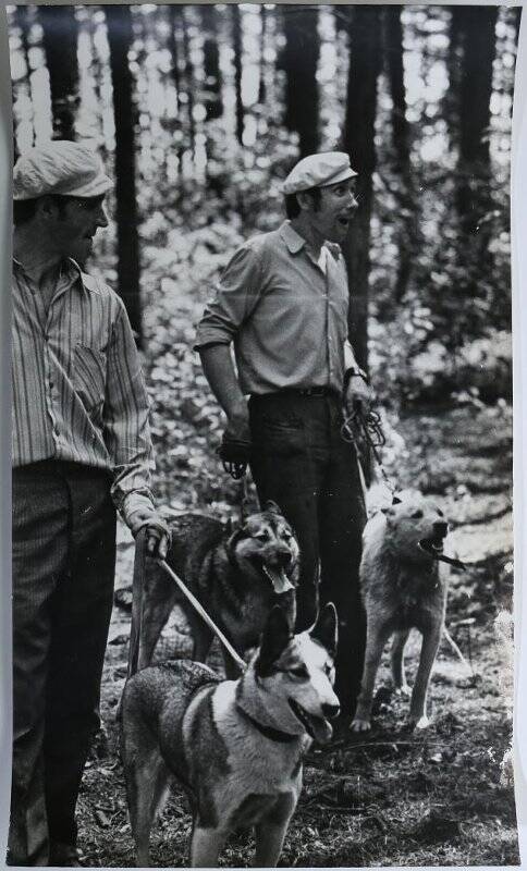 Фотография черно-белая. Двое мужчин с собаками на поводках стоят в лесу.