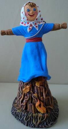 Скульптура «Сожжение  Масленицы», чучело Масленицы в голубом платье и со сковородой наперевес, водружено на разгорающийся костёр
