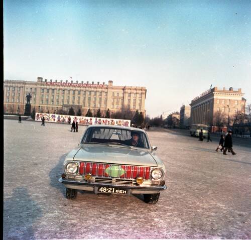 Негатив. В кадре Волга 24 стоит в центре площади Революции. На заднем плане новогодние баннеры