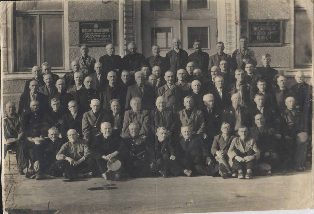 Фотография черно-белая, групповая. Изображена группа ветеранов Великой Октябрьской социалистической революции на фоне здания Горкома КПСС г. Минеральные Воды - 7 ноября 1957 год.