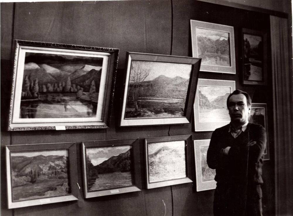 Фото ч/б событийное. На снимке в зале на стене висят картины Акимушкина В.А. он стоит на снимке справа возле своих картин.
