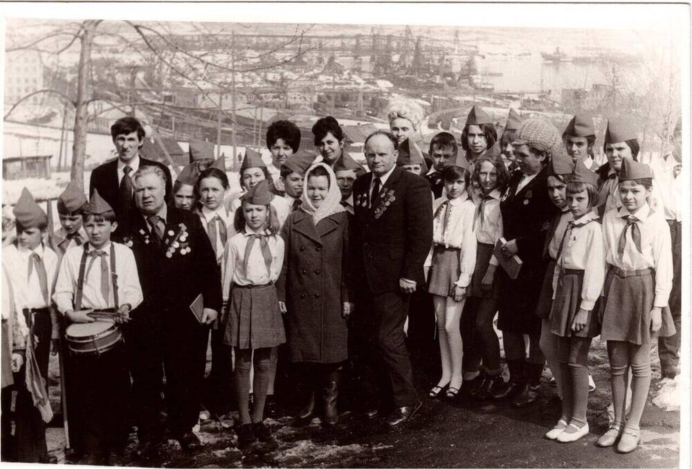 Фото ч/б событийное на фоне порта Ванино снята группа людей, состоящая из детей и взрослых. Дети - это пионеры в галстуках и парадной формы. На снимке присутствуют ветераны порта (четвертая справа Плясова Т.Д.)События 1980 года на празднике победы.