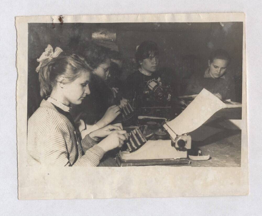 Фотография черно-белая, групповая. Изображены девочки школьного возраста, сидящие за пишущими машинками.