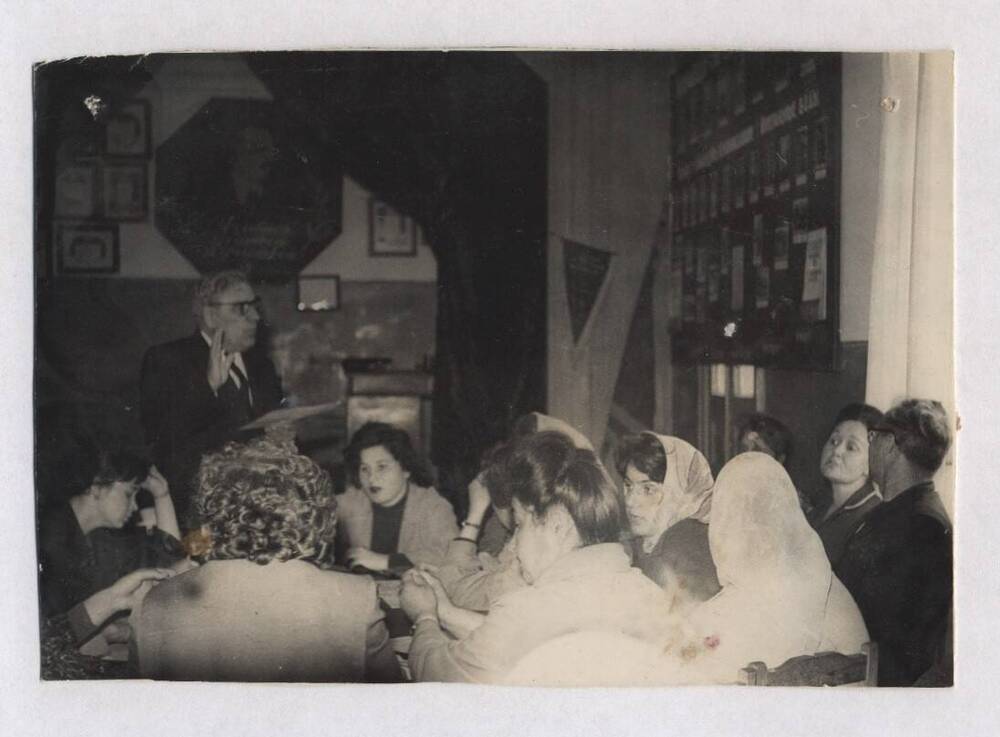 Фотография черно-белая, групповая. Изображена группа людей, сидящих за столом в пионерской комнате.