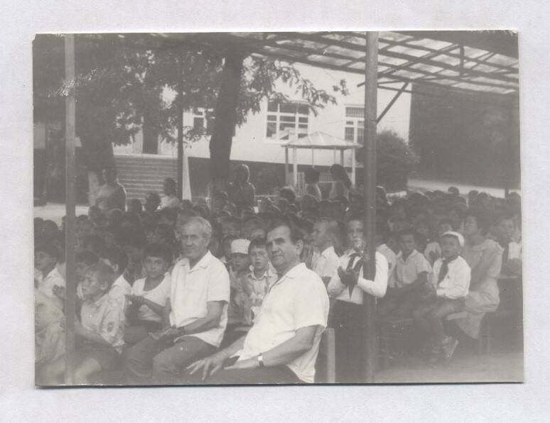 Фотография черно-белая. Изображены люди, сидящие в летнем кинотеатре. Среди зрителей много детей в пионерской форме. На краю сидит В.А. Нашивочников.