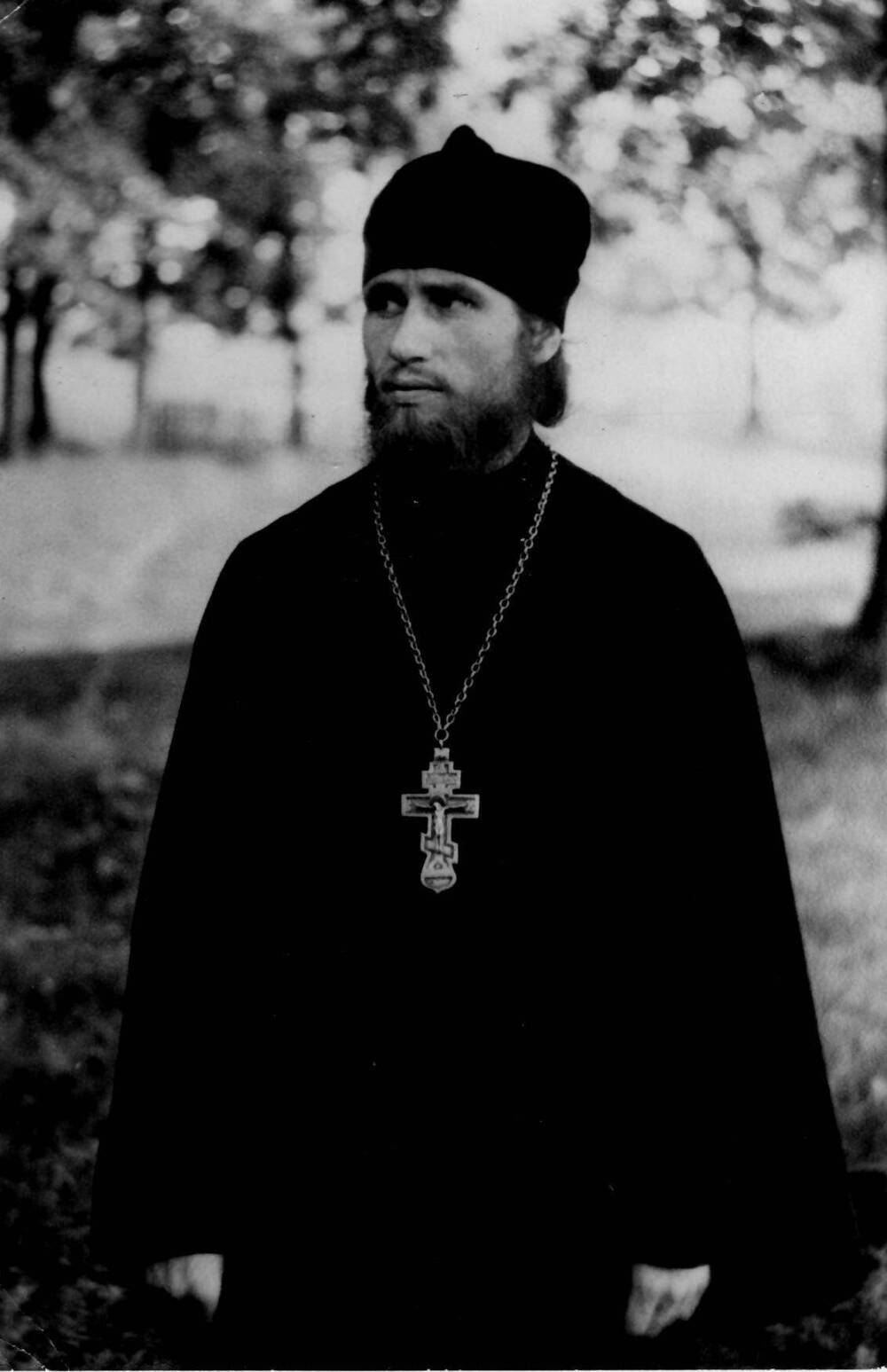 Фотография отца Геннадия в темном облачении, в скуфье с наперсным крестом на груди на фоне деревьев, 1960-1970-е гг.