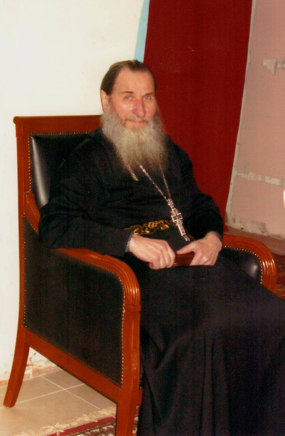 Фотография отца Геннадия в темном облачении, сидящего в кожаном кресле, с наперсным крестом и в поясе, 2011 г.