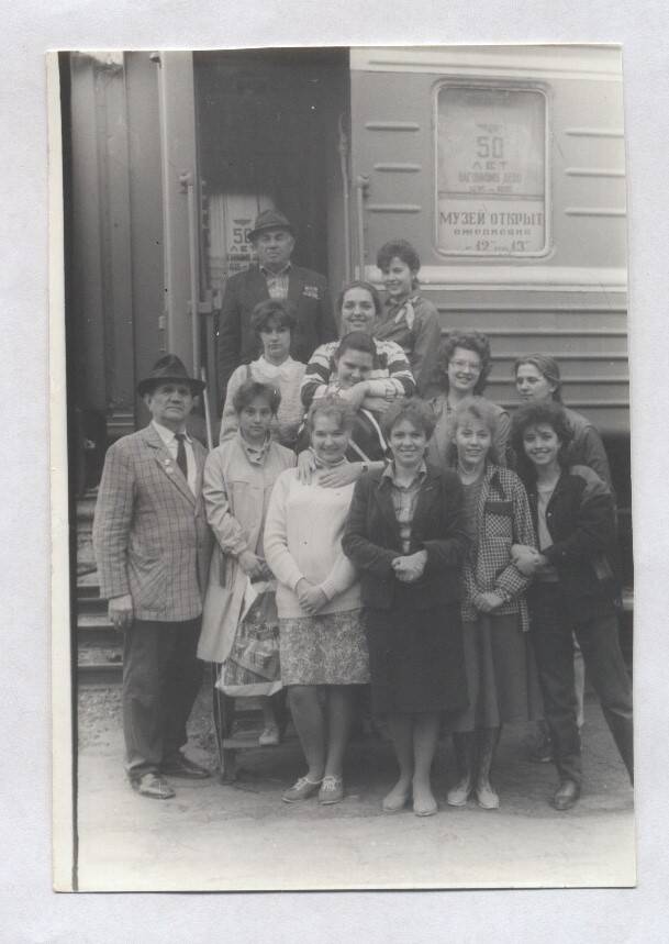 Фотография черно-белая групповая. Изображен В.А. Нашивочников, стоящий у  вагона-музея вагонного депо в кругу девушек.