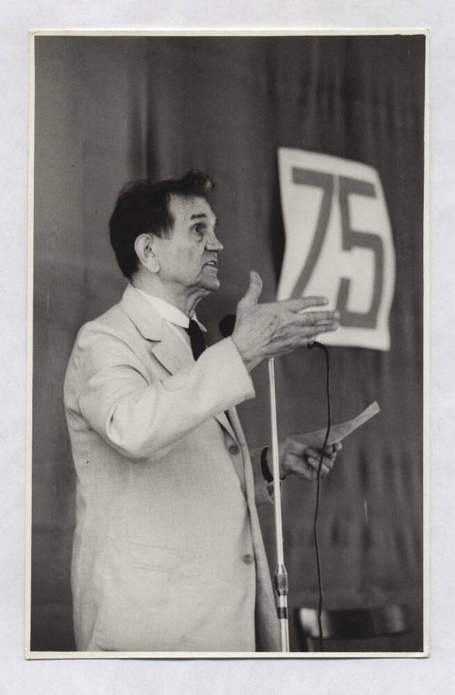 Фотография черно-белая. Изображен В.А. Нашивочников в светлом костюме, белой рубашке, черном галстуке, стоящий у микрофона с листом бумаги в левой руке.
