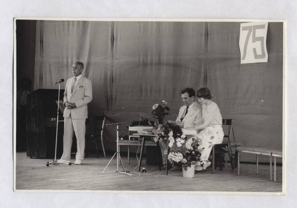 Фотография черно-белая, групповая. Изображен В.А. Нашивочников, сидящий за столом на сцене.