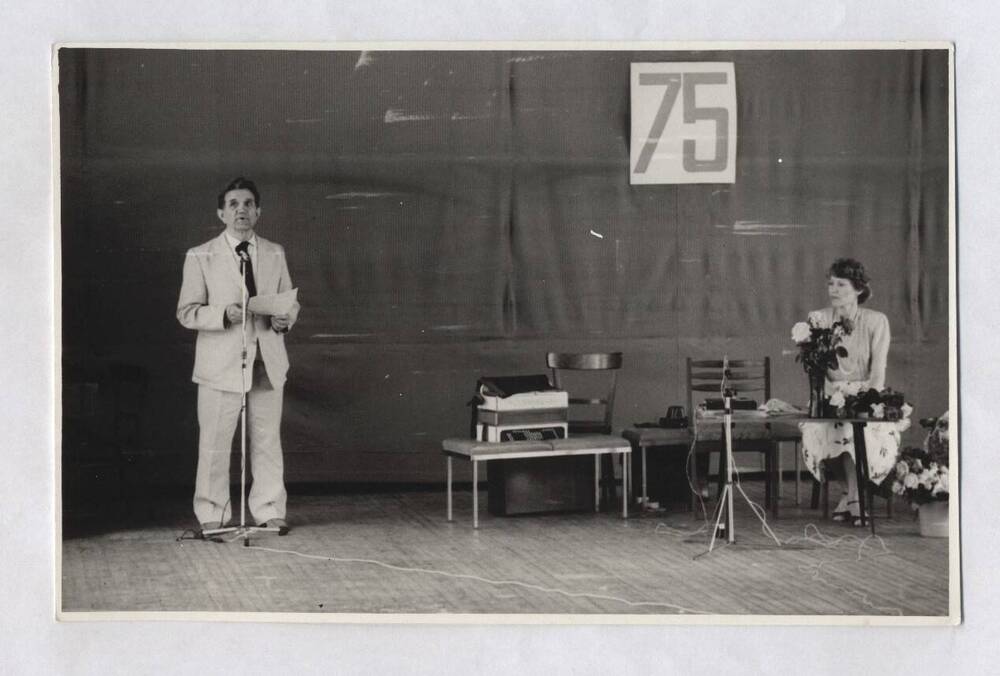 Фотография черно-белая. Изображен В.А. Нашивочников, стоящий на сцене у микрофона с листом бумаги в руках.