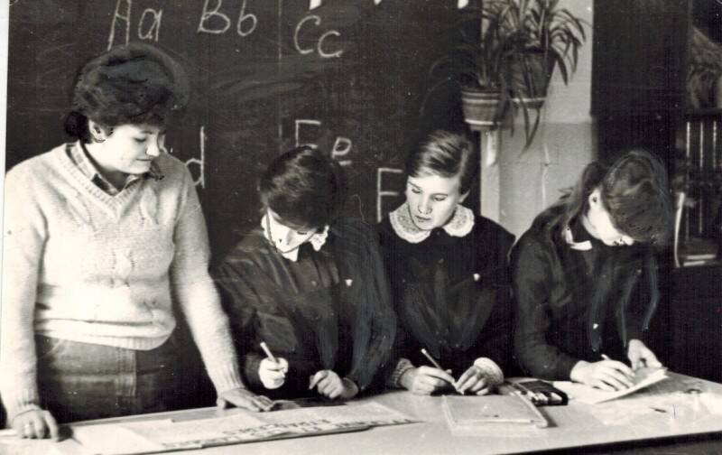 Фотография черно-белая. Сюжетная. На фотографии запечатлено четыре человека: женщина-учитель и четыре школьницы у стола.