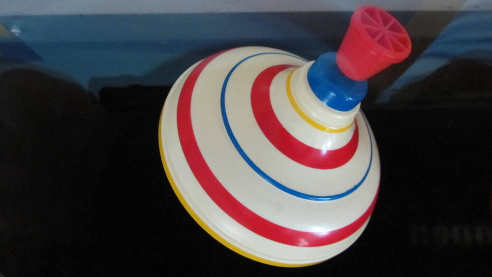 Юла - детская механическая игрушка, верхняя часть окрашена в белый цвет, декорирована цветными горизонтальными полосками разной ширины, с пластмассовой ручкой красного цвета, нижняя часть окрашена в зелёный цвет. Производитель неизвестен, 2000-е гг.