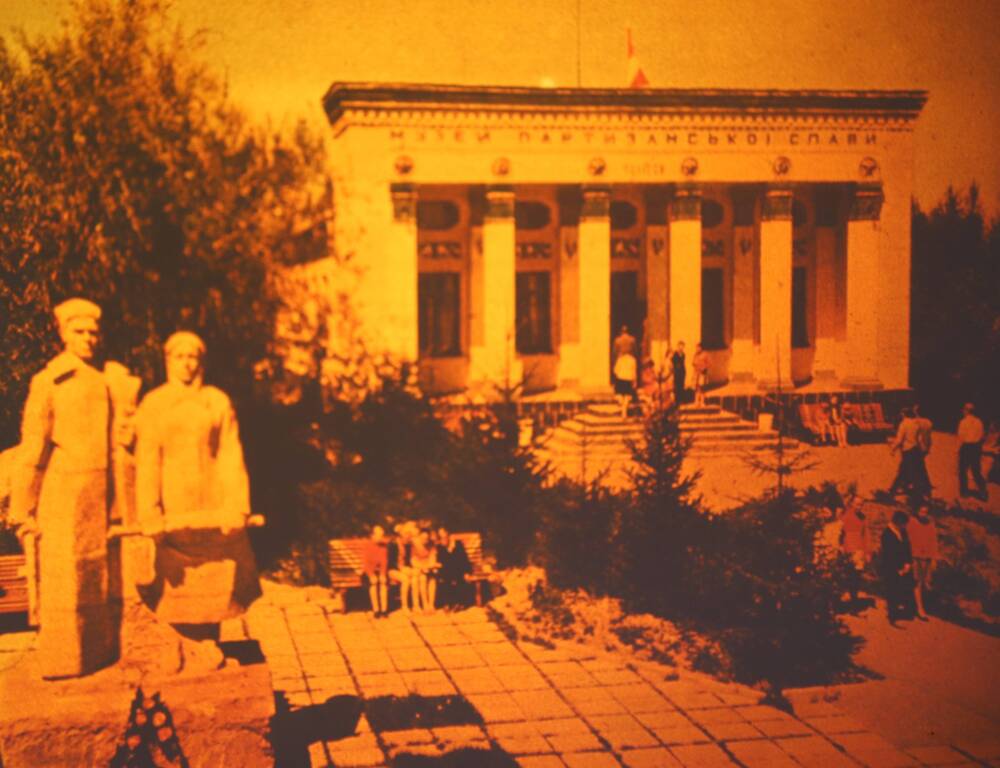 Слайд пластмассовый, где на переднем плане- на постаменте фигура мужчины и женщины с автоматами, на заднем плане- здание, на котором высечен текст-«музей боевой славы» на украинском языке.