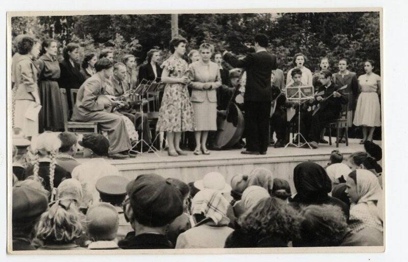 Фото. На сцене выступает коллектив художественной самодеятельности, спиной к зрителю на сцене стоит дирижер в темном костюме; две молодые женщины исполняют песню; позади них полукругом расположились музыканты со струнными инструментами, перед ними на высоких подставках установлены ноты; позади сцены - сплошная зелень высоких деревьев; на первом плане - головы зрителей, смотрящих на сцену. 1950-е г.г.; г. Зарайск.