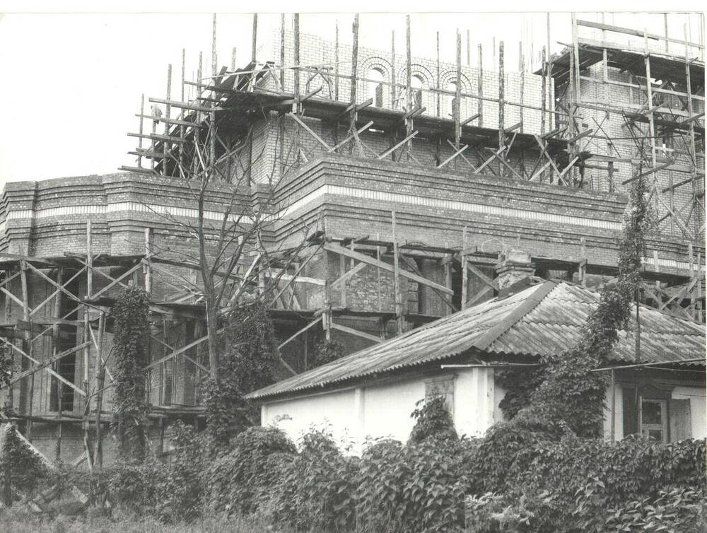 Фотография черно-белая.  Общий вид строительства нового храма в строительных лесах. На переднем плане соседний жилой дом с вьющимися кустарниками.
