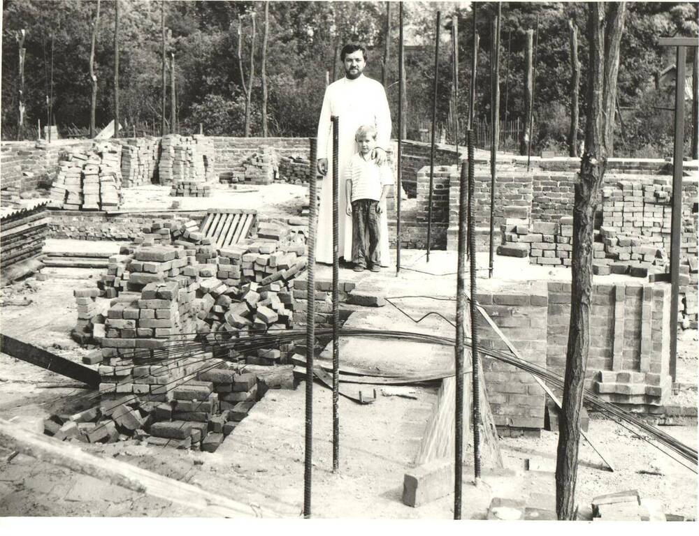 Фотография черно-белая. Г.М. Самойленко с сыном Михаилом на строительстве нового храма. За спиной алтарная часть храма, деревья сада.