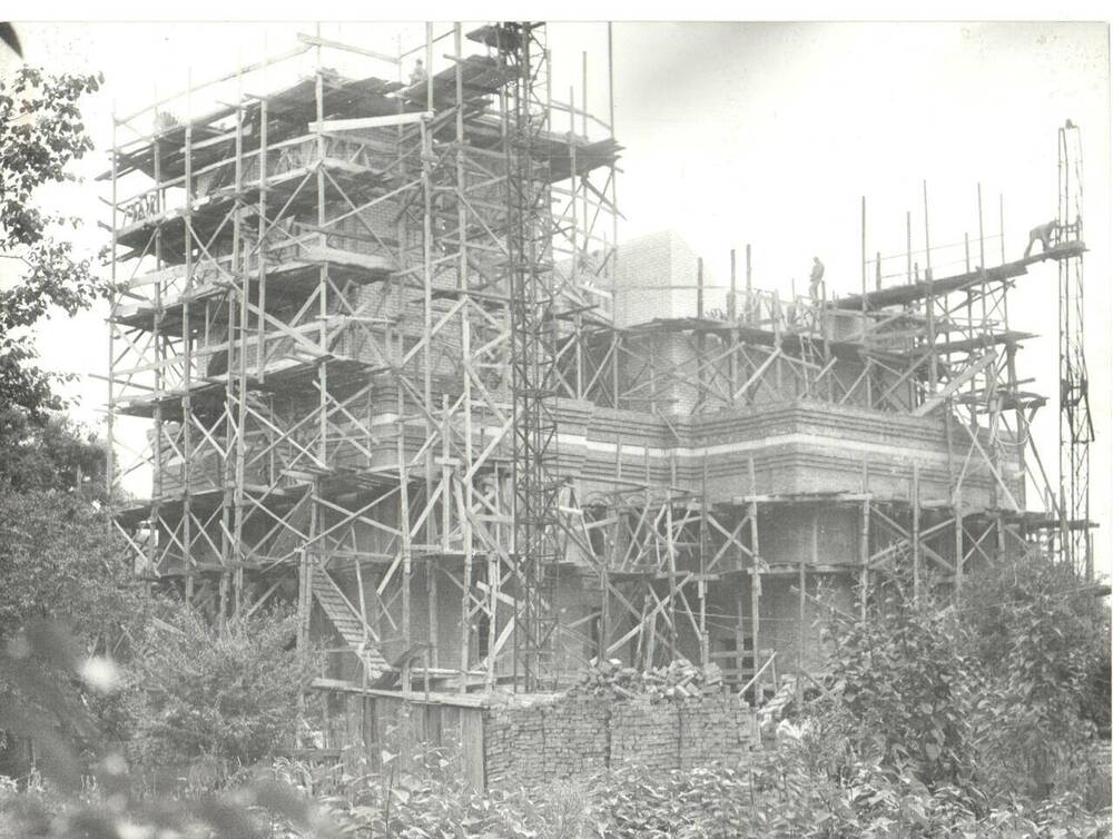 Фотография черно-белая.  Общий вид строительства нового храма. Строительство колокольни  с северо-западной стороны нового строения в строительных лесах. На переднем плане деревья, кустарники, кирпич.