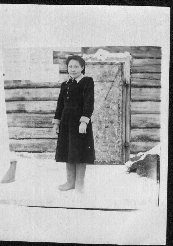 Фотография черно-белая. Девушка неизвестная в полный рост в темном платье с белым воротничком и манжетами, в серых валенках, на улице снег, на фоне деревянного здания.