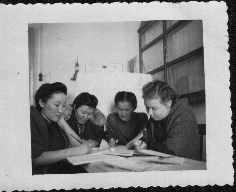 Фотография черно-белая групповая (4 неизвестные девушки). Сидят за столом на занятии на фоне книжной полки.