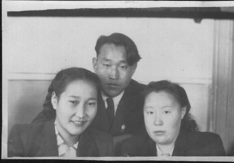 Фотография черно-белая групповая. Неизвестные 2 девушки, сидят, парень стоит посередине на заднем плане.