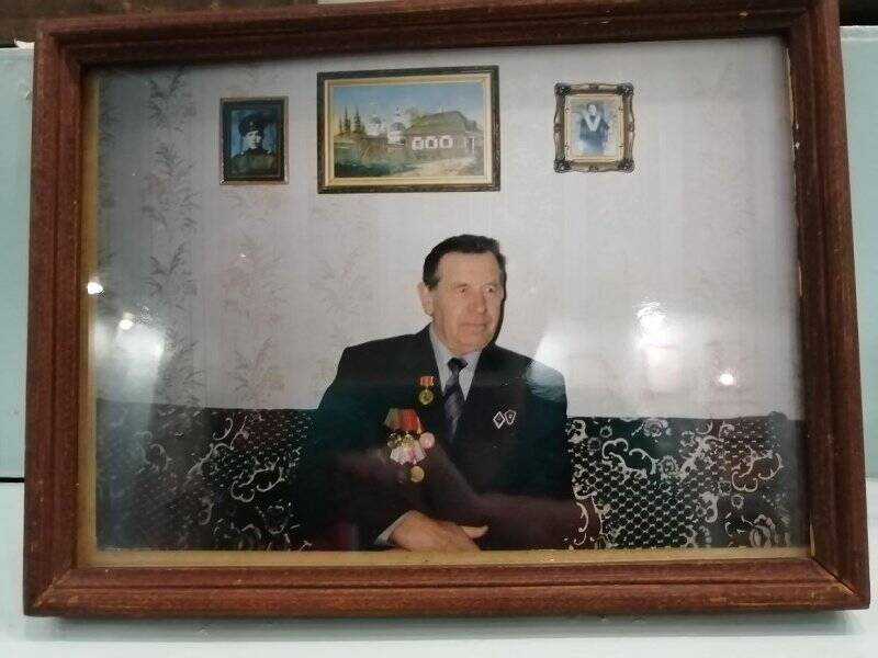 Фотография в рамке. Фото цветное  «Петр Николаевич Астраханцев» . Сидит на диване, на стене две фотографии и картина дома. В деревянной рамке 24*18