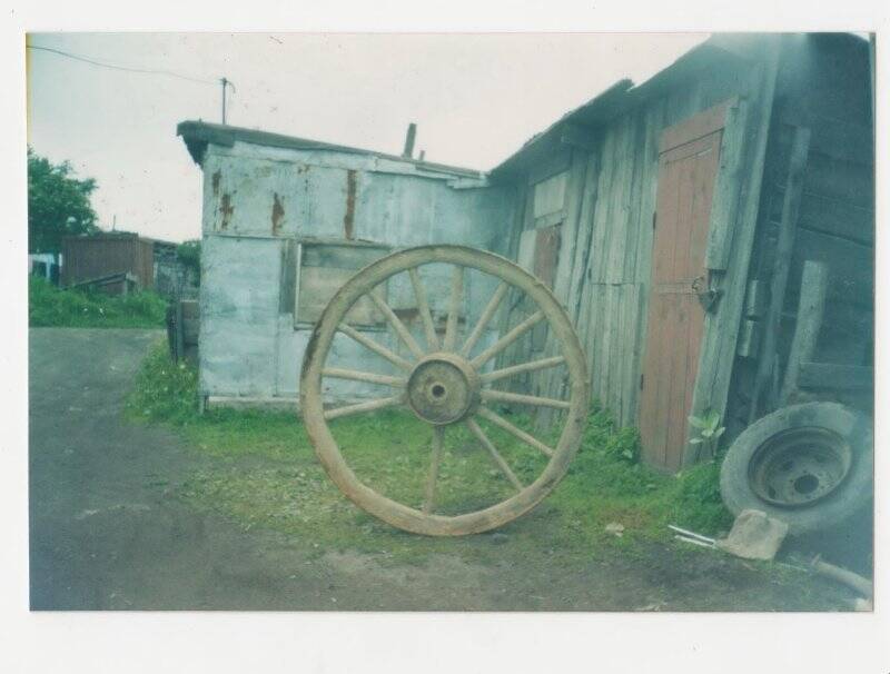 Фотография. Оригинал. Деревянное колесо от повозки или телеги (предположительно японское) на фоне хозяйственных построек на улице Южно-Сахалинской.
