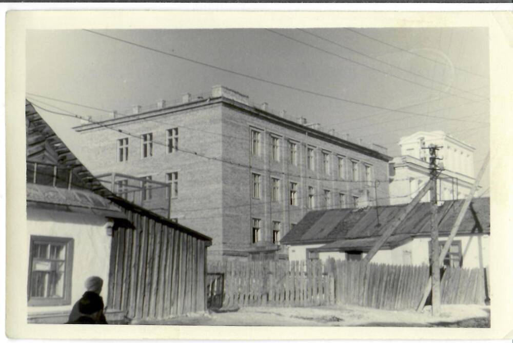 Фотография черно-белая, глянцевая печать. На фото: трехэтажное здание в окружении частных домов. На обороте машинописный текст: «Пристройка к горному техникуму на 2.1У.62г. г. Магадан», г. Магадан, 1962 г.
