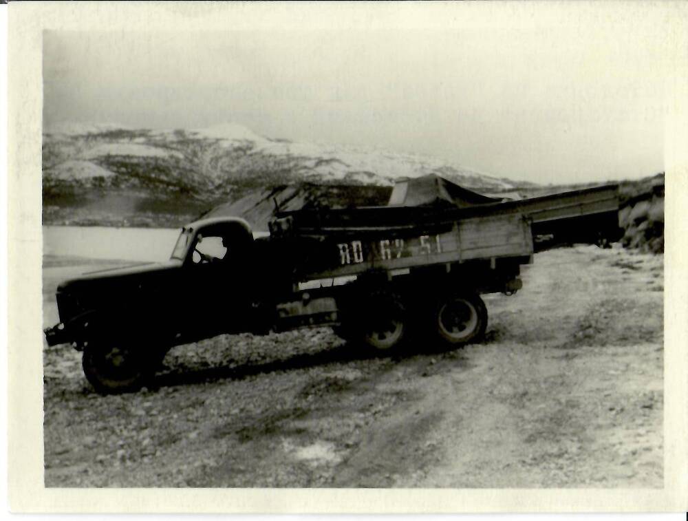 Фотография черно-белая, глянцевая печать. На фото: грузовой автомобиль с мотолодкой в кузове на берегу водоема в окружении сопок. На борту кузова написан номер белой краской [ПО 62-51], г. Магадан, 1961 г.