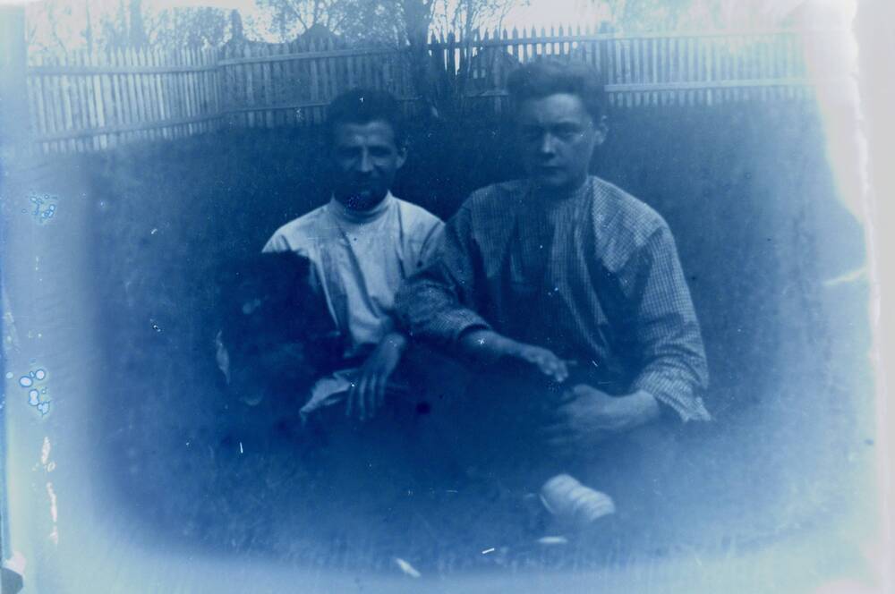 Негатив. Двое мужчин изображены на улице сидя на траве на фоне деревянного забора