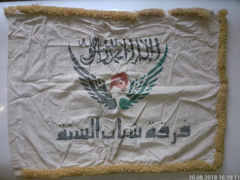 Флаг белый с арабскими надписями и жёлтой бахромой.