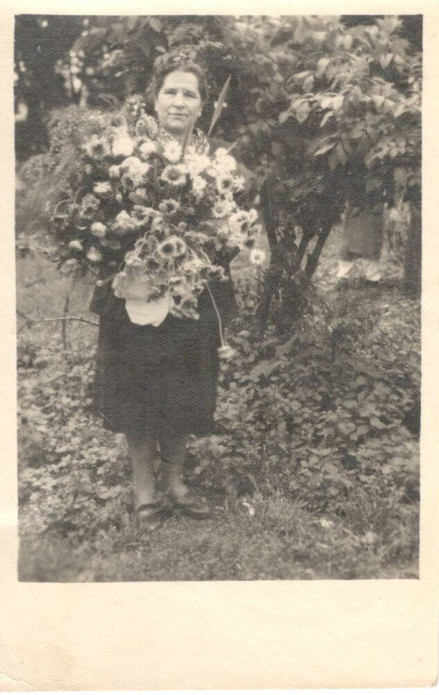 Фотография черно-белая, в рост.  В парке, на фоне  кустарника с листвой и густо растущей травой под ногами ,  с огромным букетом цветов в руках стоит Мария Андреевна Попова.