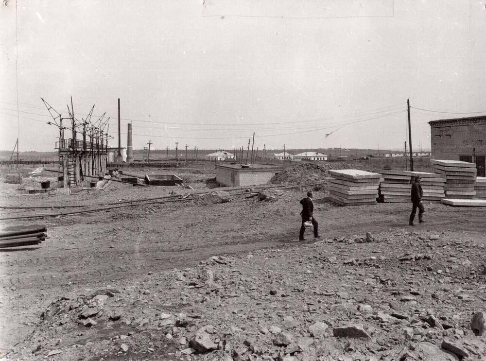 Фото ч/б документальное.Строительство нефтебазы порта Ванино 1970 г. На снимке площадка, где лежат строительные блоки, имеются арматурные постройки, на здании лежат трубы, есть кирпичные постройки. Вдали жилые одноэтажные здания. По дороге идут двое мужчин