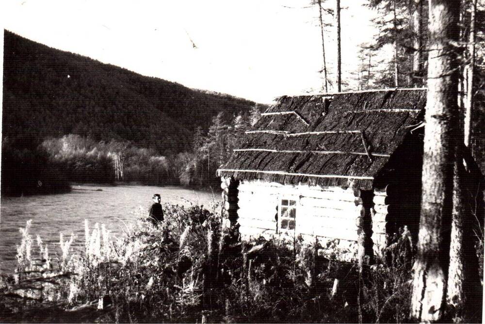 Фото ч/б документальное. На снимке крупным планом один из первых домов в Уська- Орчское, он сделан из бревен. Имеется небольшое окно. Рядом с домом стоит мужчина. Рядом течет река Тумнин.