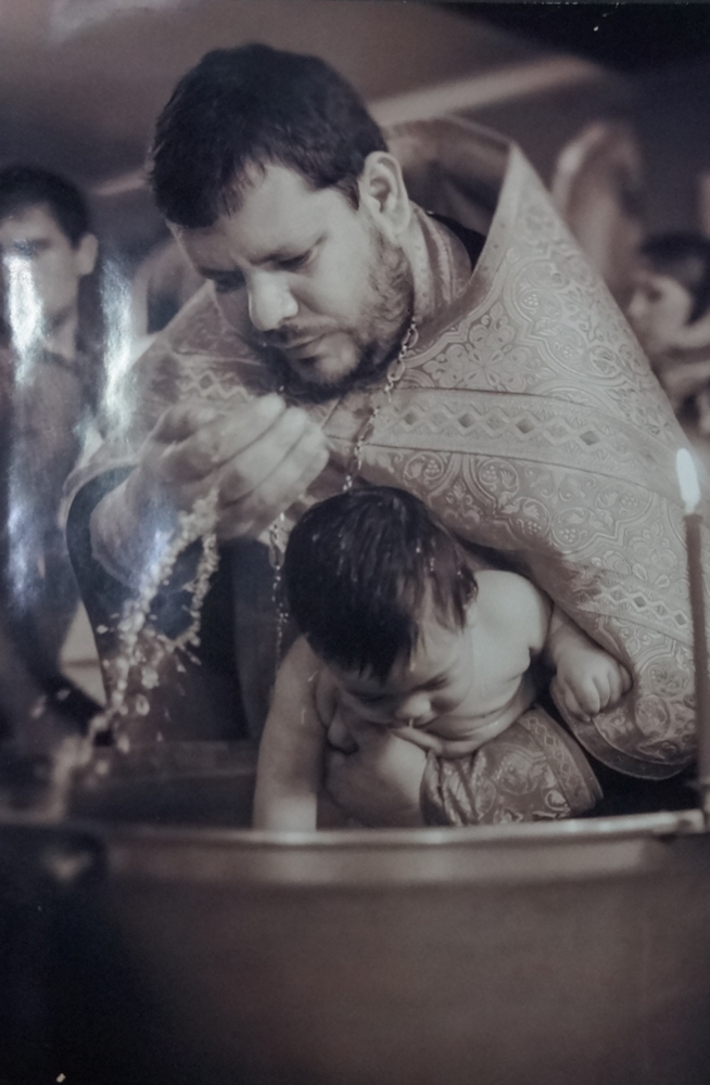 Фотография черно-белая с изображением священнослужителя, совершающего обряд крещения.