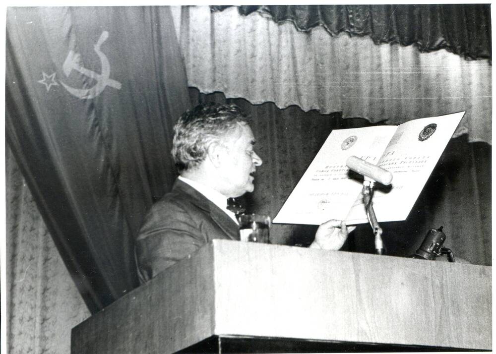 Фото ч/б. П.С. Плешаков за трибуной зачитывает Грамоту. 1980-е гг.