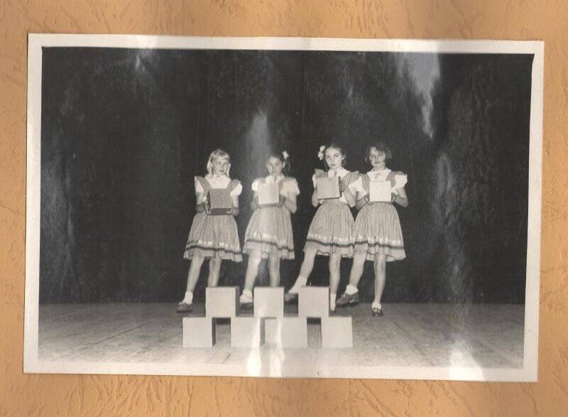 Фотография ч/б, групповая,  вертикальная, наклеена на лист картона в альбоме. На сцене четыре девочки в цветастых сарафанах (платьях?) длиной выше колена. Девочки в руках держат кубики. Перед девочками на сцене восемь кубиков пирамидой в два ряда.  ХХв.,70-80-е годы.