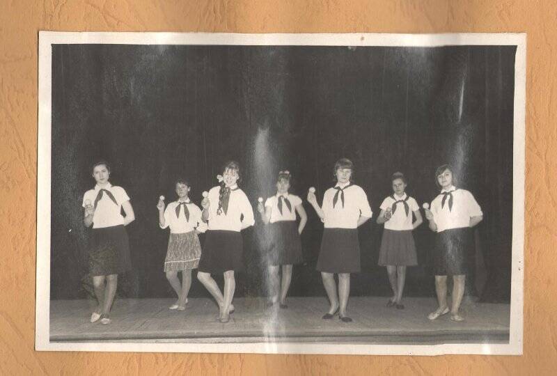Фотография ч/б, групповая, наклеена на лист картона в альбоме. На сцене семь танцующих девочек в пионерских галстуках, светлых блузках, темных юбках длинной выше колена. У второй девочки слева юбка пестрая. В правой руке у каждой девочки погремушка (?). ХХв.,70-80-е годы.