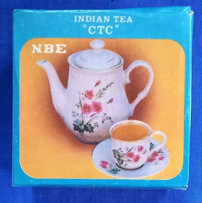 Коробка чайная «INDIAN TEA CTC NBE» картонная, голубая, глянцевая, запечатанная, чай упакован внутри. Коробка объемная - многогранник с с шестью гранями. На  квадратных сторонах коробки напечатаны цветные изображения. На одной- заварочный чайник с чашкой и блюдцем, на другой - женщины в национальной индийской одежде с коробами за спиной, собирающие листья чая на плантации. Масса нетто 250 г. Производство – Кочин, Индия.