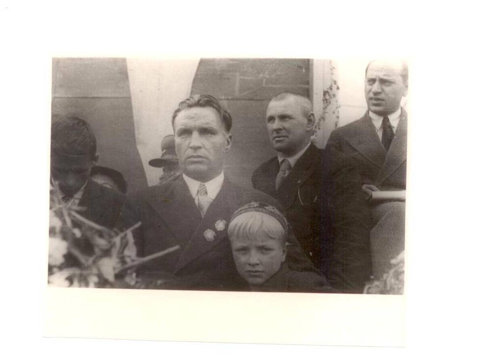 Фото. В. П. Чкалов в штатском костюме без головного убора стоит на трибуне в пос. Василево, рядом с ним его сын Игорь.