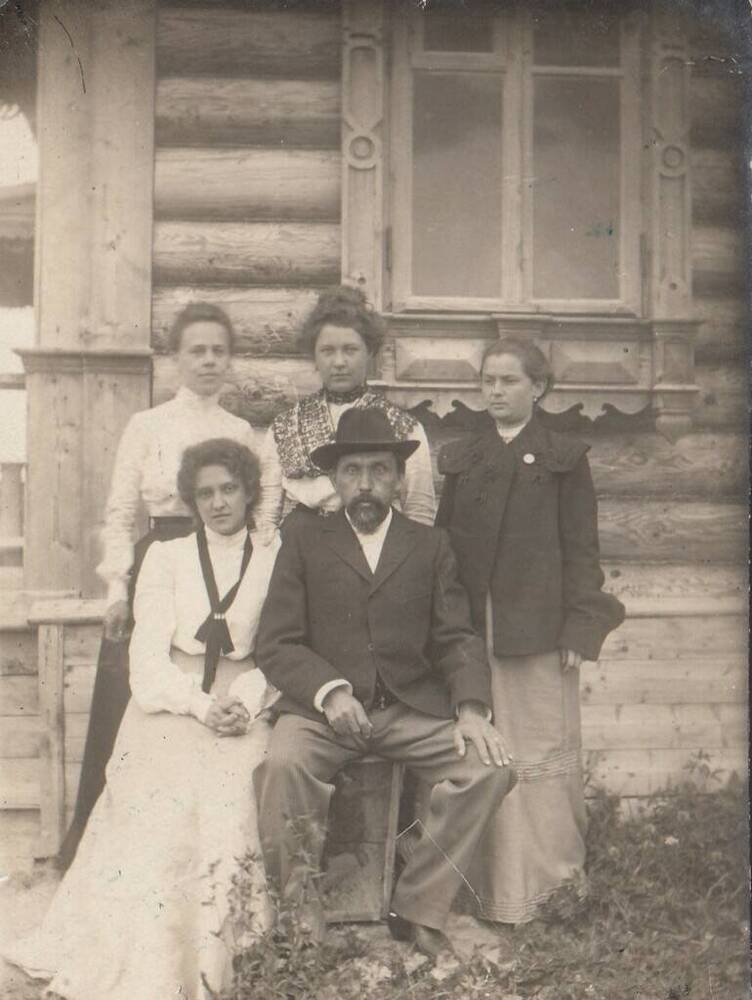Фотография групповая - Г.Н.Прозрителев с молодыми женщинами на фоне бревенчатого дома; Прозрителев Г.Н. и Загорская В.Г. сидят на деревянном ящике, за ними стоят три молодые женщины. Место съемок и время неизвестны.