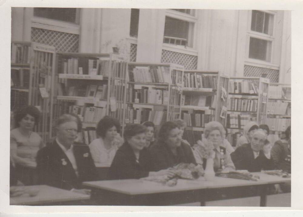 Фотография, черно-белая, групповая Заики-на Анна Абрамовна-участница Великой Отечественной войны 1941-1945 гг.  в библиотеке. Сидит за столом (в первом ряду 4-я слева). Рядом с ней за столом слева 3 чело-века, справа один. За спиной семь человек. На заднем плане стеллажи с книгами.