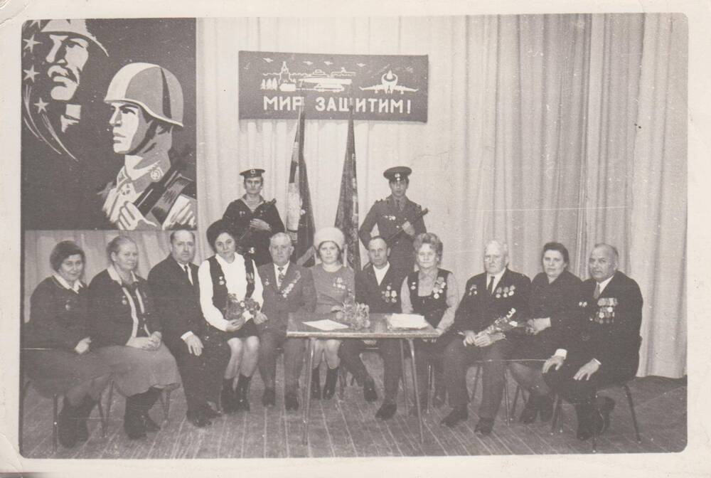Фотография, черно-белая, групповая Заики-на Анна Абрамовна-участница Великой Отечественной войны1941-1945 гг.  сидит за журнальным столиком (восьмая слева) Одета в светлую блузу темный сарафан. На груди справа и слева ордена и медали. По обе стороны от нее сидят мужчины и жен-щины, награжденные орденами и медалями. За их спинами у флагов стоят два солдата в военной форме. На стене слева висит кар-тина с изображением солдат с оружием в руках, в центре схематично изображен Кремль, с надписью: «Мир